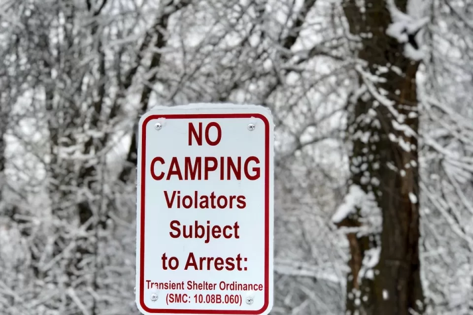 White sign with No Camping - Violators Subject to Arrest and municipal code in red text with snowy trees in background.
